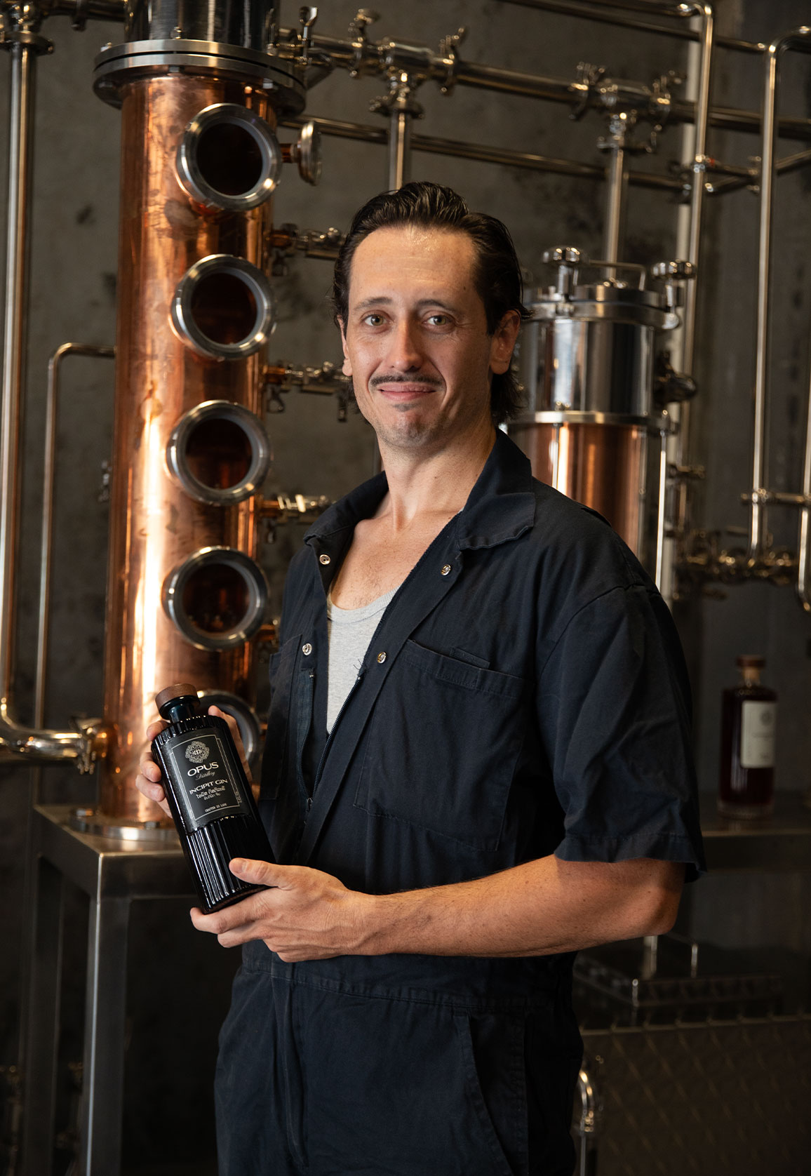 Charles Leglise, founder of Opus Distillery, holding an Incipit Gin bottle in front of copper stills in Laos