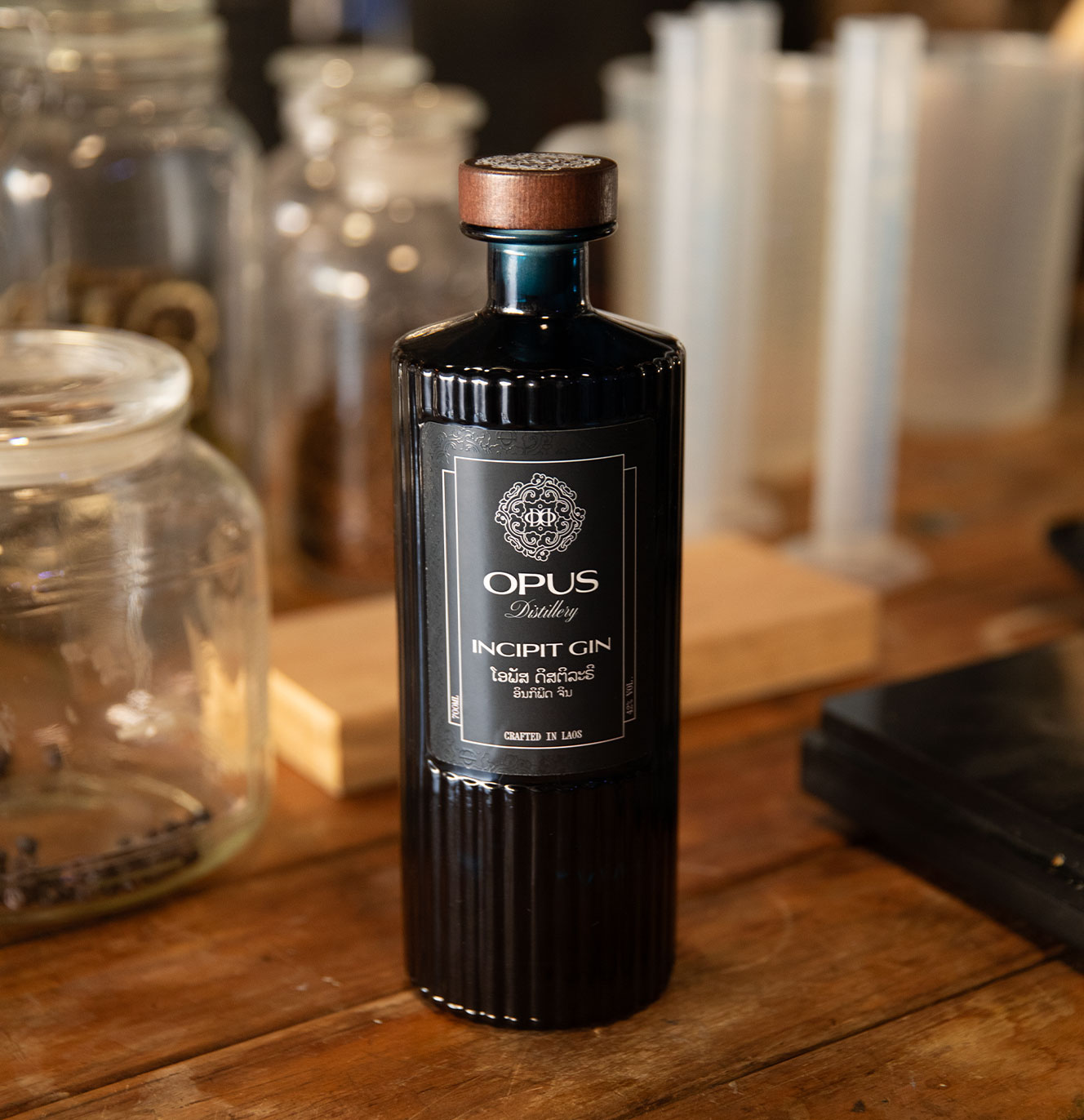 Opus Distillery Incipit Gin bottle on a wooden workbench surrounded by glass jars and measuring cylinders in the distillery